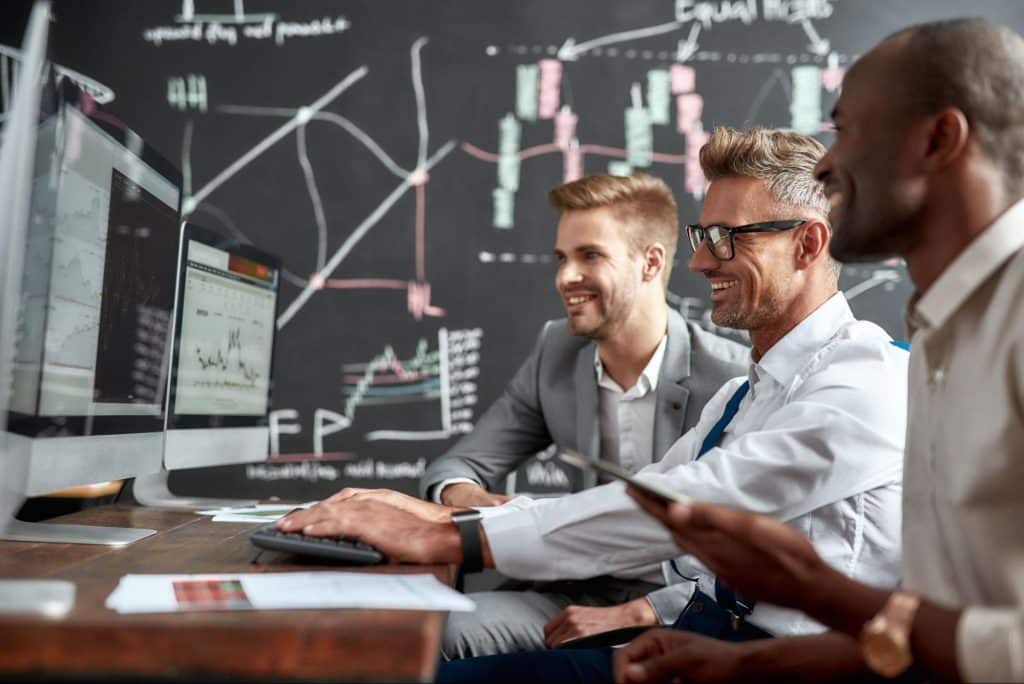 Três homens em escritório olham sorridentes para computadores enquanto analisam dados da empresa.