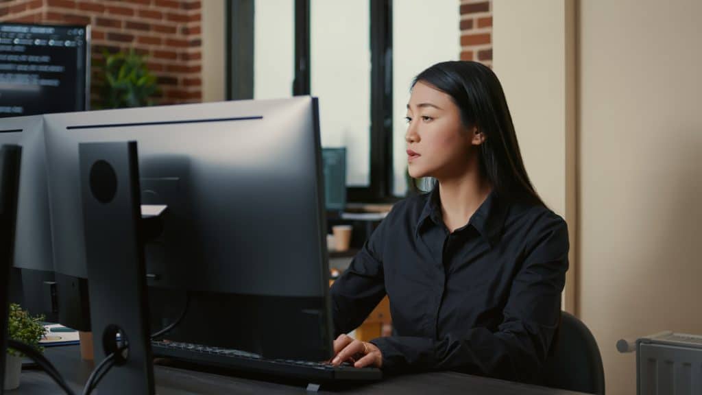 mulher asiática sentada em um escritório, olhando para as telas de computador a sua frente.
