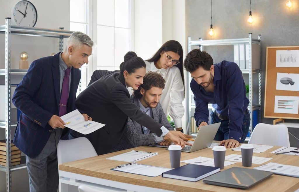 Grupo de pessoas diversas em escritório se aproximam de uma grande mesa e encaram dados em um notebook.