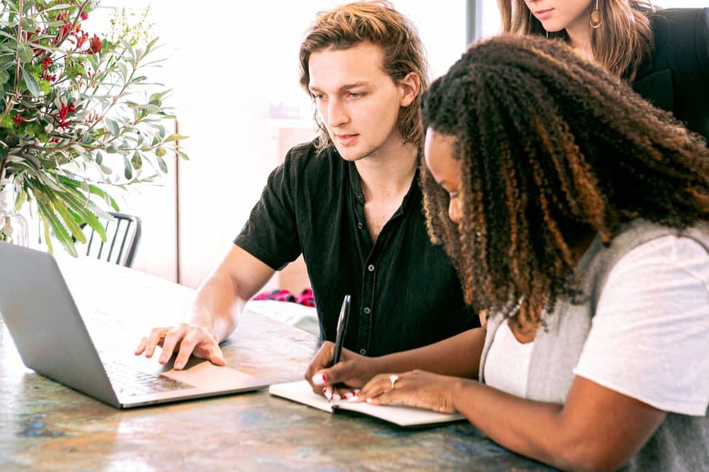 Grupo diverso em mesa de escritório com notebook à frente. Um homem branco encara a tela, enquanto uma mulher negra ao seu lado faz anotações em um caderno.