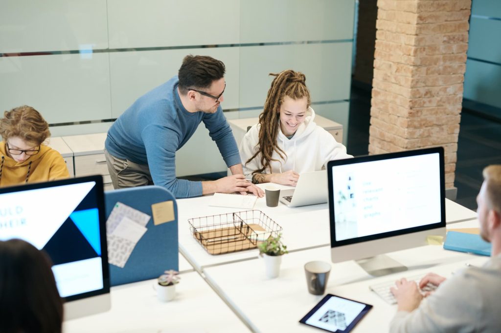 Escritório com grupo diverso sentado de frente para diferentes computadores. Ao centro, um homem de óculos conversa com uma mulher de dreadlocks, que sorri. 