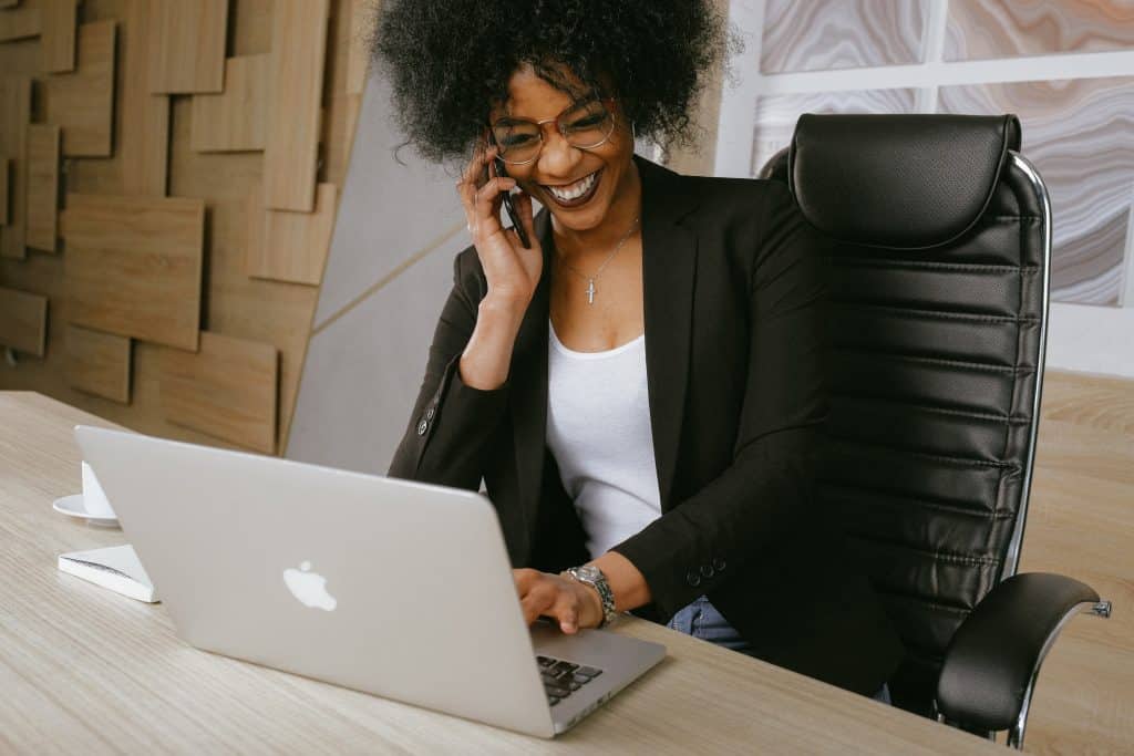 Mulher negra de óculos sentada em uma cadeira sorri com um celular na orelha enquanto mexe em um notebook à mesa.