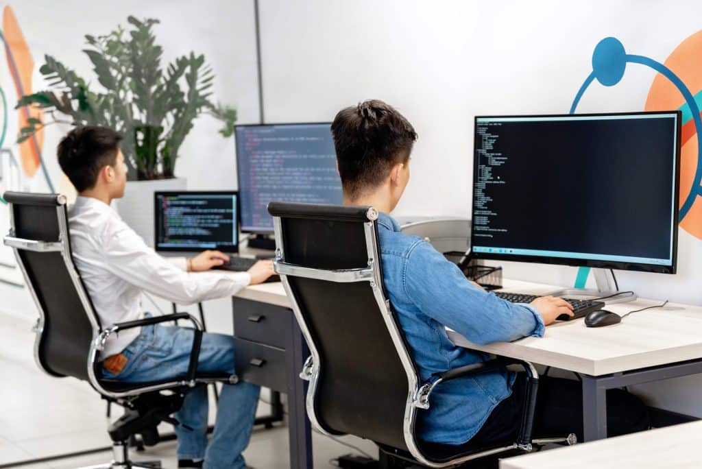 dois homens sentados de frente para computadores em um escritório, programando.