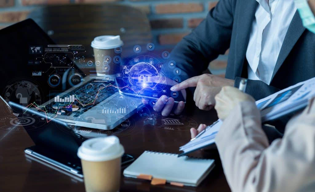 Duas pessoas em uma mesa de escritório, com celulares, cadernos e copos de café ao redor. Do celular, um holograma com ícones de finanças e gráficos é projetado.