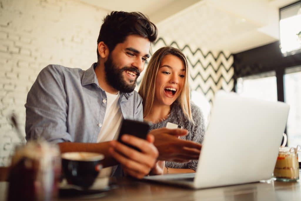 Homem com celular na mão encara notebook sobre a mesa, com mulher loira ao lado com um cartão de banco na mão.