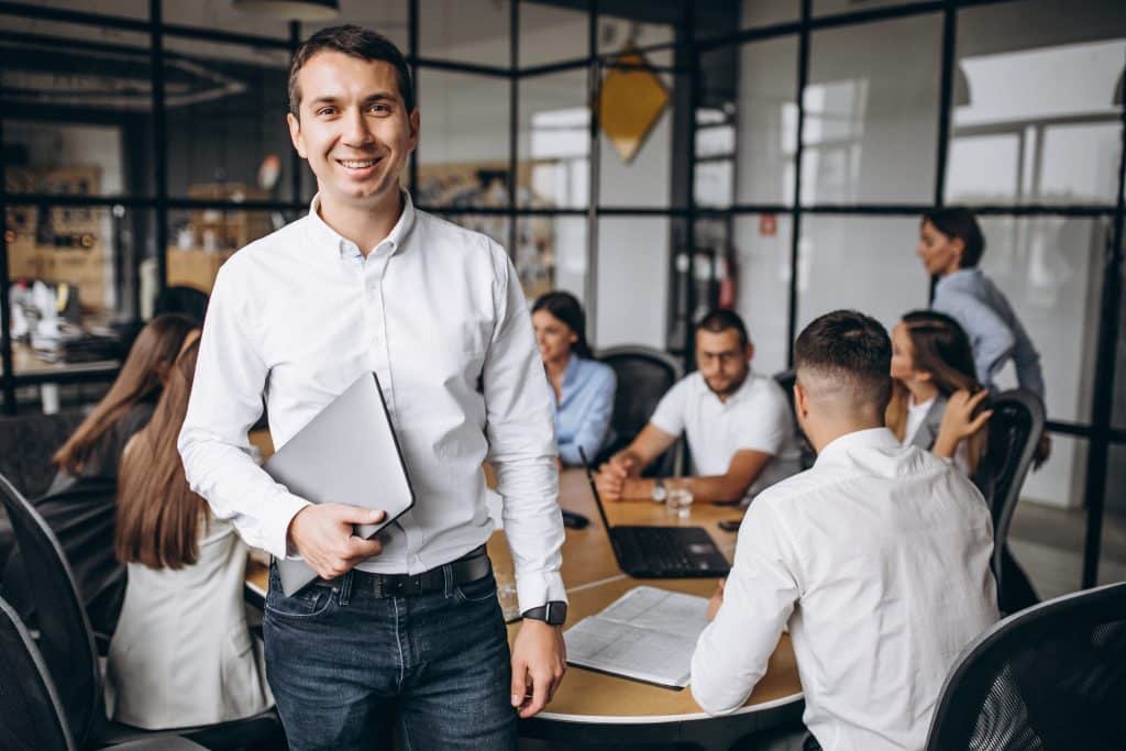 Profissional da tecnologia sorridente segurando um notebook fechado, enquanto ao fundo, um grupo de profissionais estão sentados ao redor de uma mesa, em reunião.