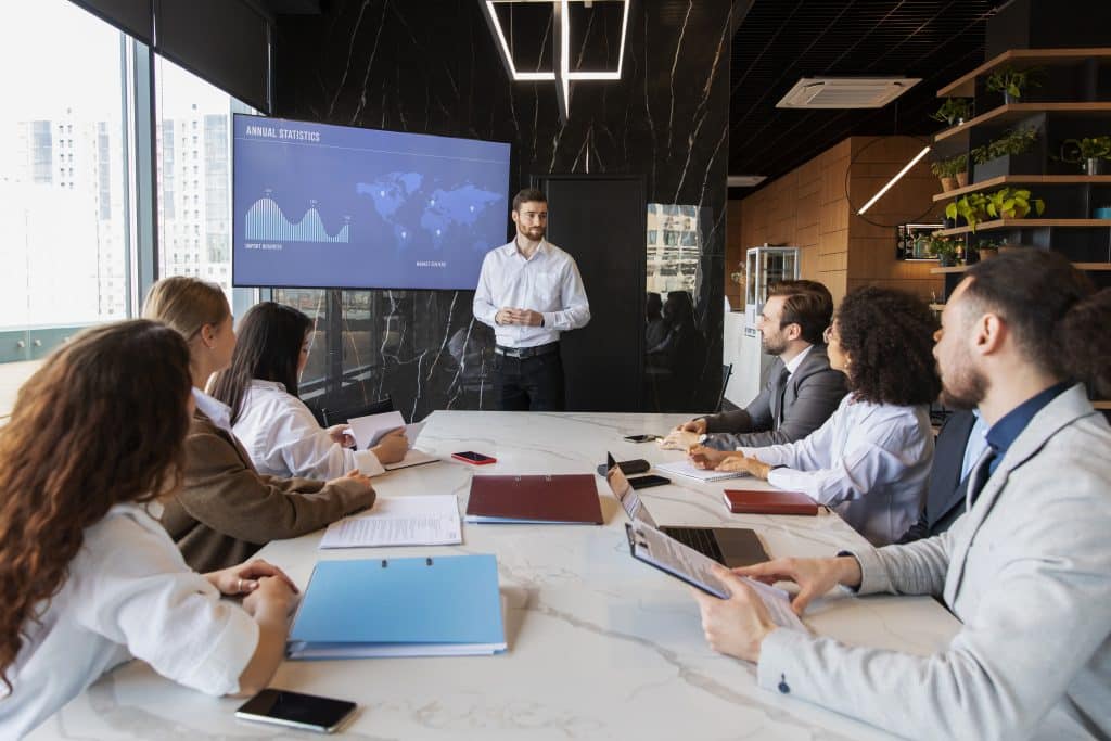 Profissionais da tecnologia em uma reunião, enquanto seis deles estão sentados ao redor de uma mesa, um deles está apresentando gráficos e mapas em uma tela.